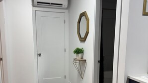 A hallway with a white door, an air conditioning unit, a decorative mirror, a small plant, and a dark-colored cabinet. -     Island Escape | Cozy 2BR Suite Steps from the Shore (Parksville)