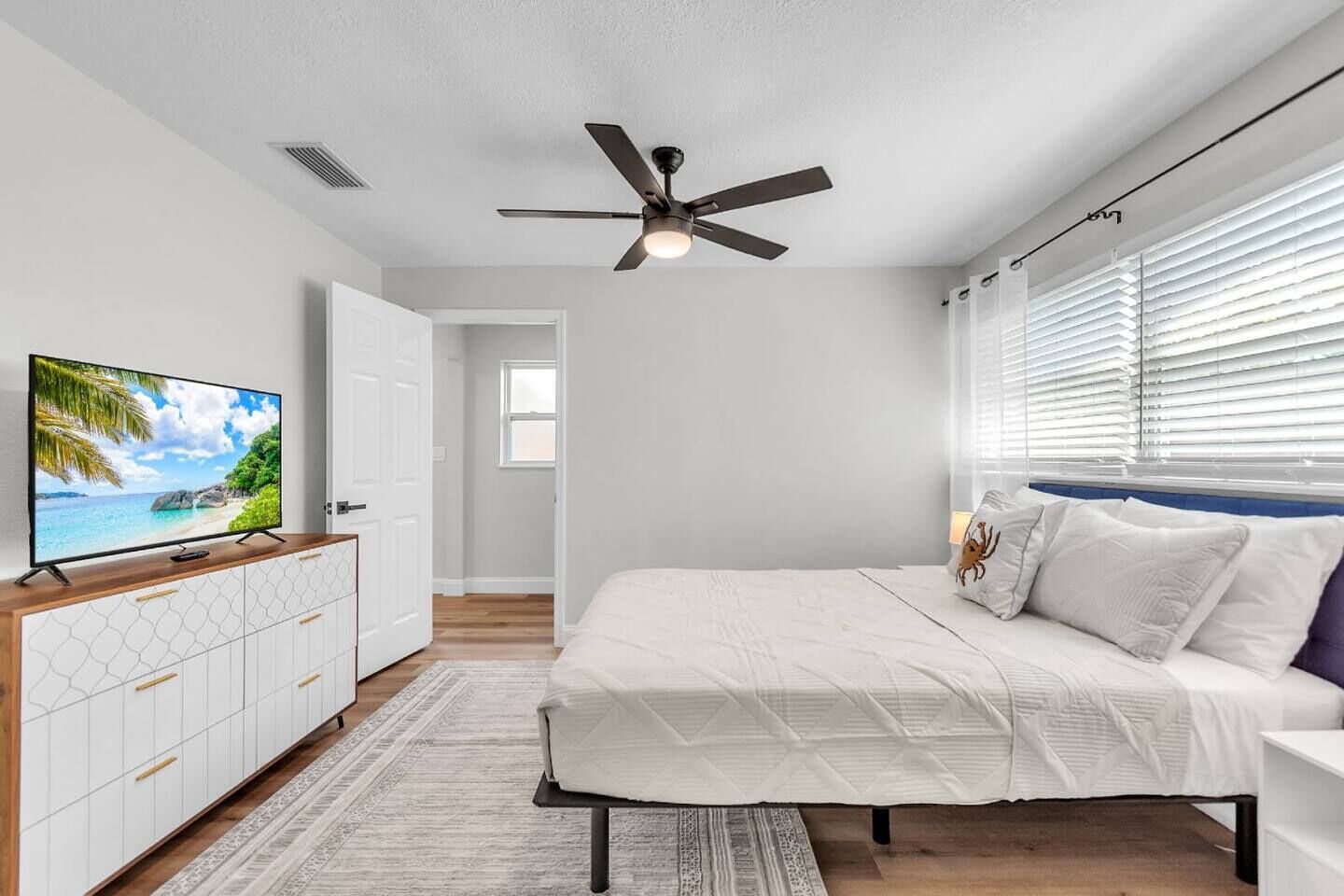 Ein Schlafzimmer mit einem Bett, einem Fernseher auf einer Kommode, einem Deckenventilator und einem Fenster mit Jalousien.