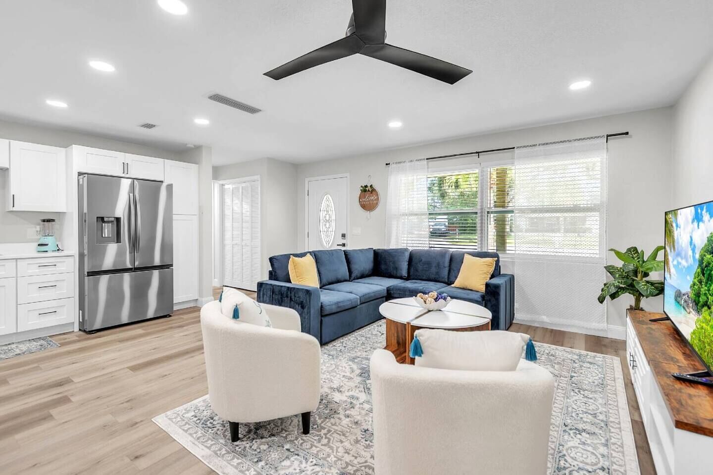 A modern living room with a sectional sofa, a coffee table, and a kitchen area with a stainless steel refrigerator.