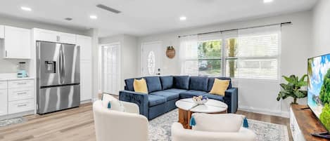 A modern living room with a sectional sofa, a coffee table, and a kitchen area with a stainless steel refrigerator.