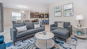 A modern living room with a grey sofa, ottoman, and round coffee table. There is a kitchen in the background with a dining area. - Cozy 6BR Home,Sleeps 16,Near Disney & Universal FL (Four Corners)
