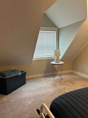 A small, sloped ceiling room with a window, a table with a lamp, a black sofa, and a bed.
