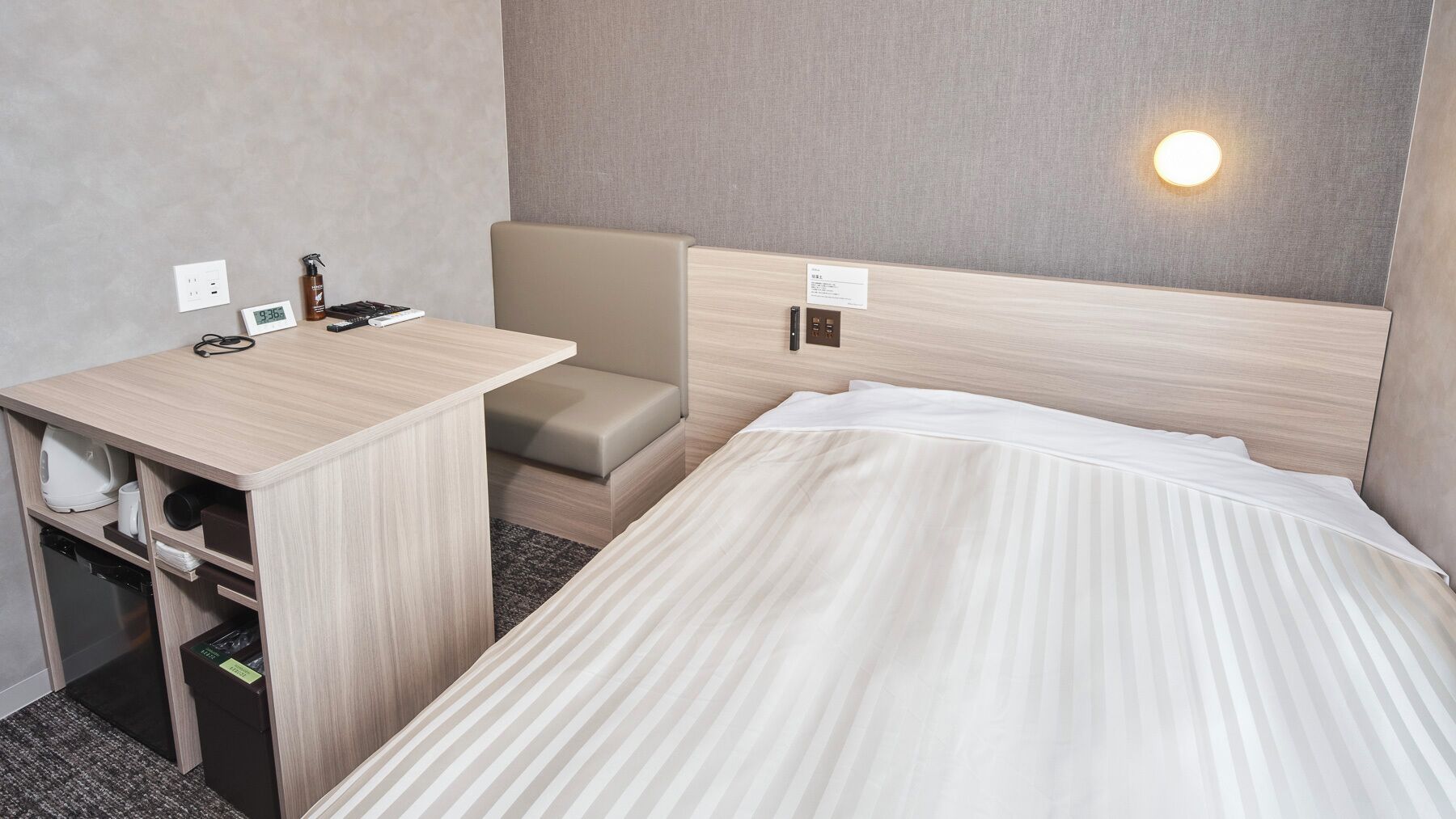 A hotel room with a bed, a desk with a chair, a small sofa, and a wall-mounted light.