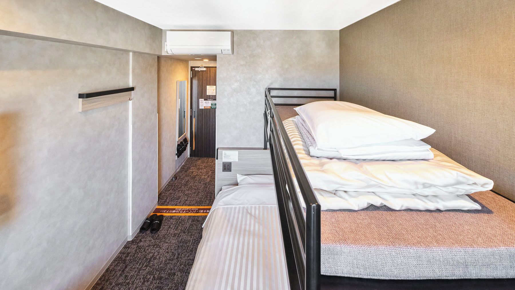 Habitación de hotel estrecha con una cama litera, suelo alfombrado y un pasillo que conduce a un baño.
