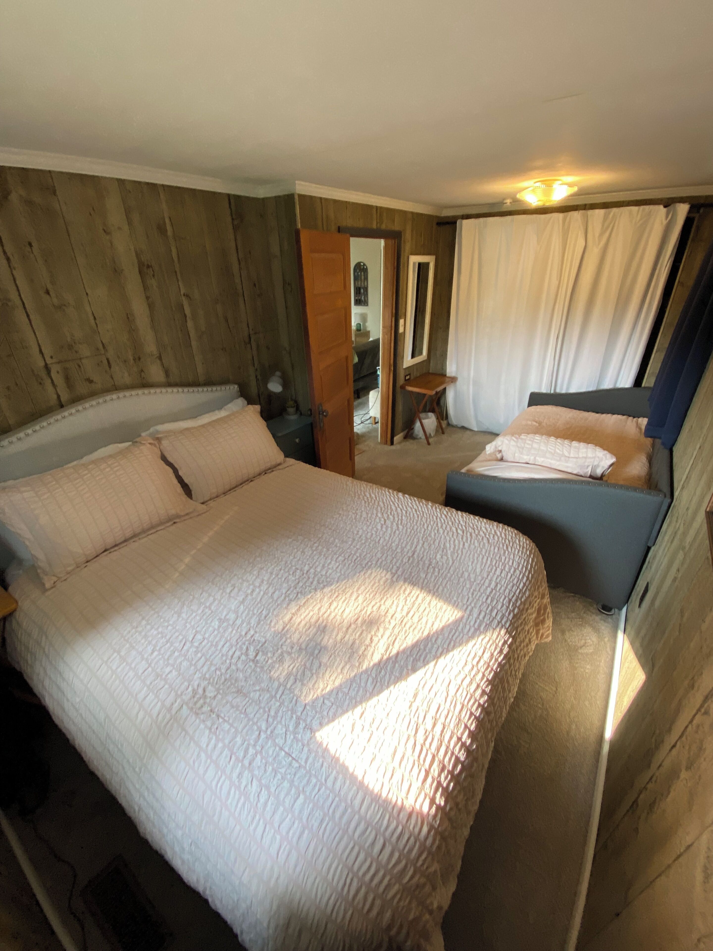 A bedroom with a large bed, a smaller bed, a wooden wall, a mirror, and a window with curtains.