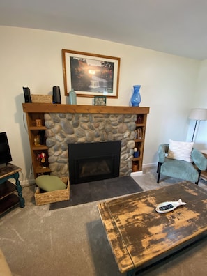 Una sala de estar con chimenea de piedra, un cuadro enmarcado encima, un jarrón azul y un sillón color aguamarina.