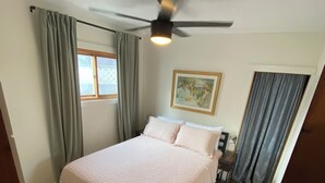 Une chambre à coucher avec un lit, un ventilateur de plafond, une fenêtre avec des rideaux et un tableau au mur.