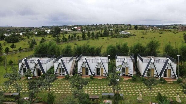A row of white, triangular-roofed cabins with balconies, set in a green landscape with a distant view of houses.