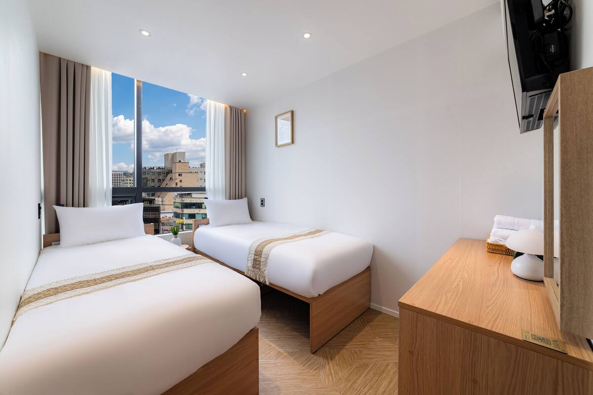 Quarto de hotel com duas camas, uma escrivaninha de madeira, televisão e uma grande janela com vista para a cidade.