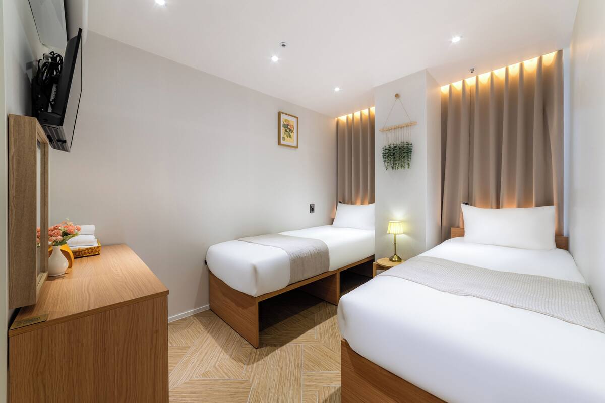 Quarto de hotel com duas camas, uma escrivaninha de madeira, televisão e uma pequena planta.