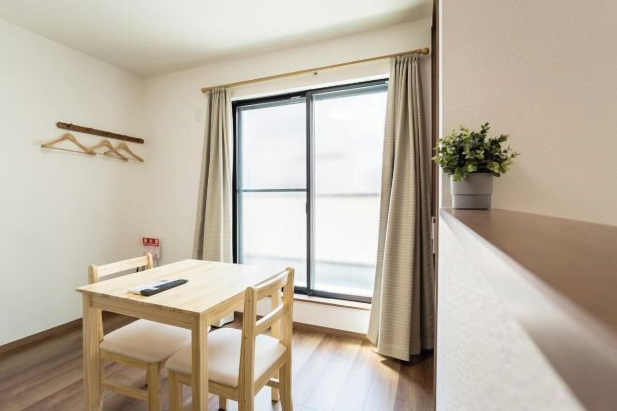 A room with a wooden table and chairs, a window with curtains, a plant on a shelf, and a coat rack with hangers.