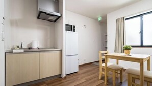 A modern kitchen with a white refrigerator, a microwave, and a coffee maker on the counter. There is a wooden dining table with two chairs and a small potted plant. - HANASTAY UMI (Osaka)