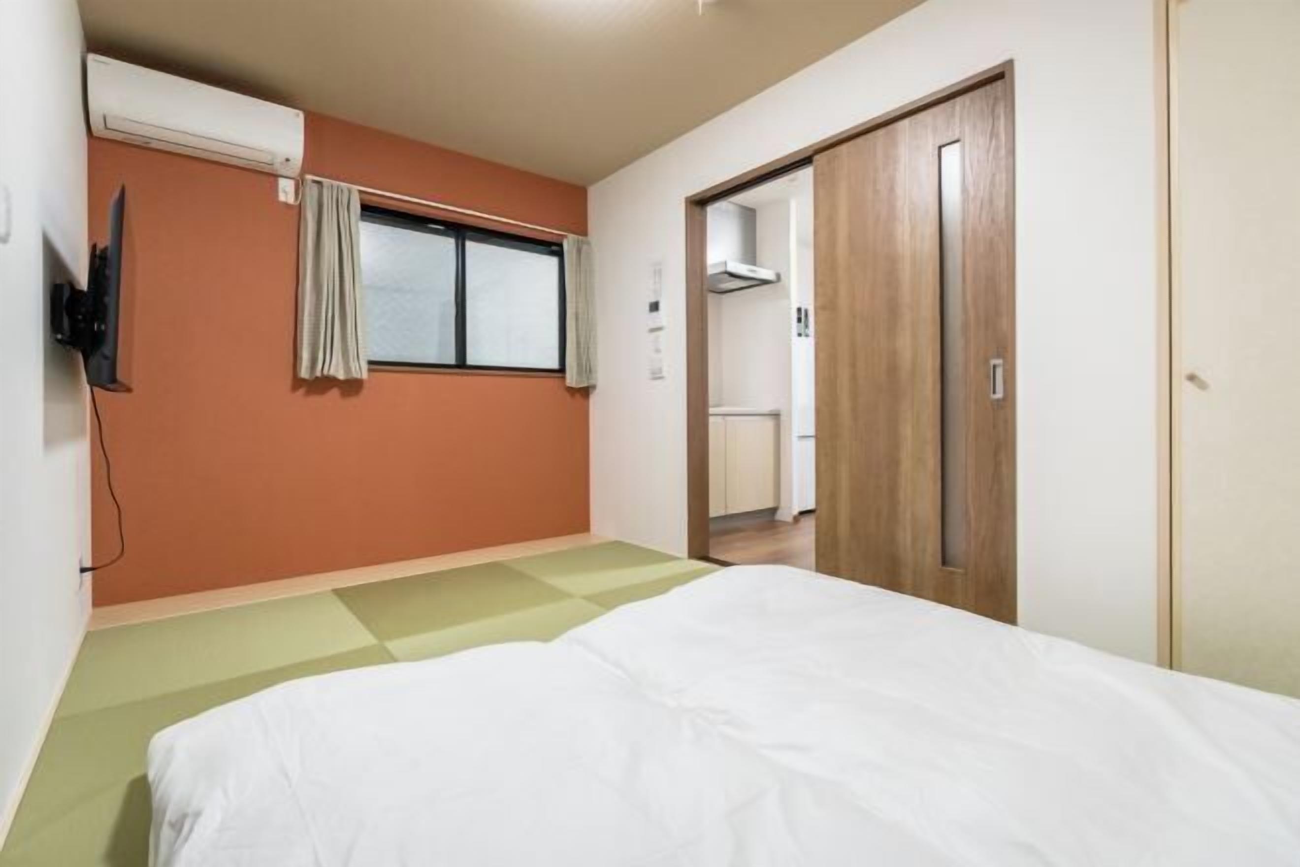 A bedroom with a bed, a television mounted on the wall, a window with curtains, and a wooden door leading to a bathroom.