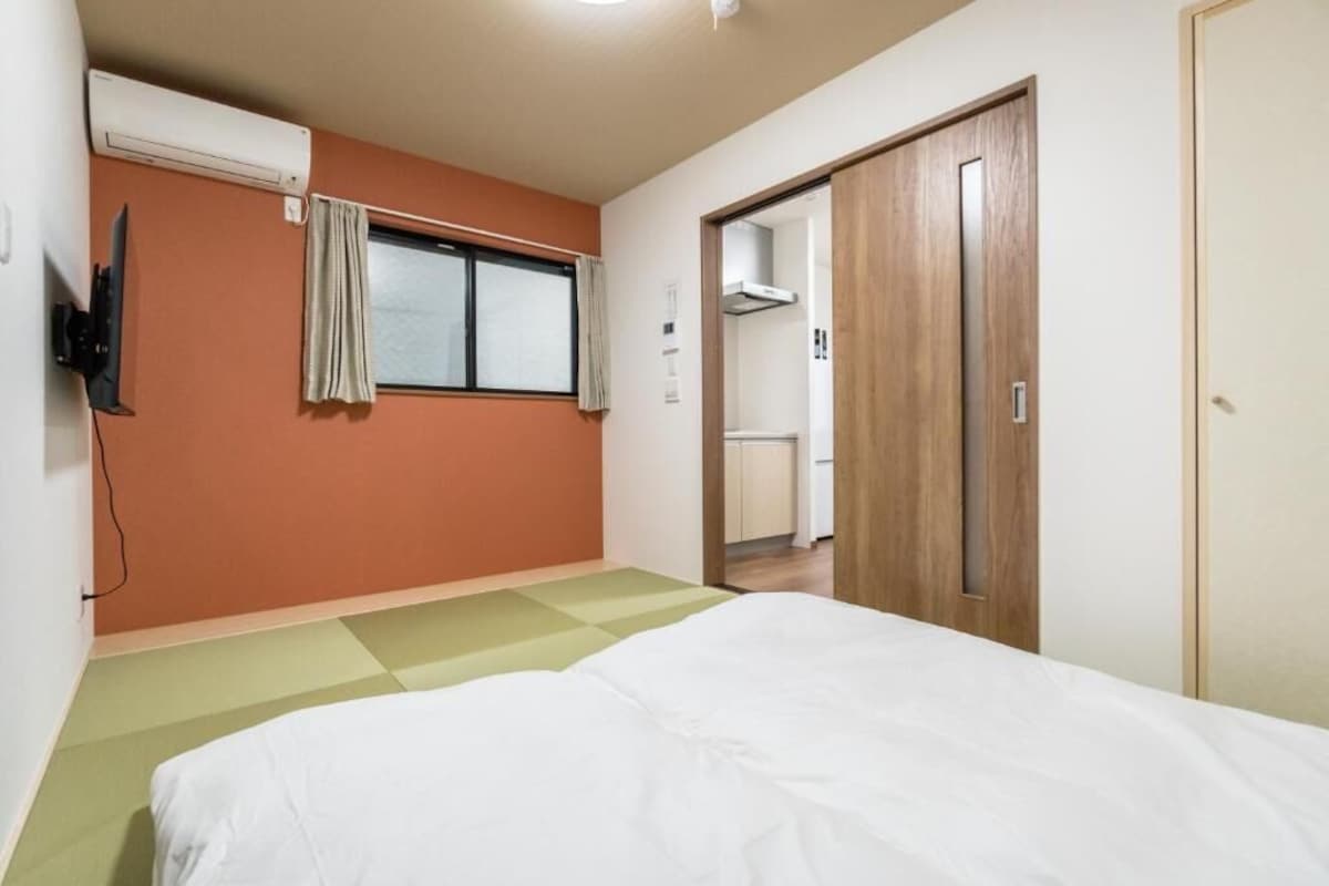 A bedroom with a bed, a window with curtains, an air conditioning unit, a wooden wardrobe, and a small kitchenette area.