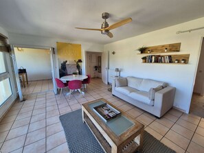 A living room with a white sofa, a coffee table, a ceiling fan, a dining table with red chairs, and a bookshelf. - Sea view, 4 sleeps, all on foot (ST MANDRIER SUR MER)