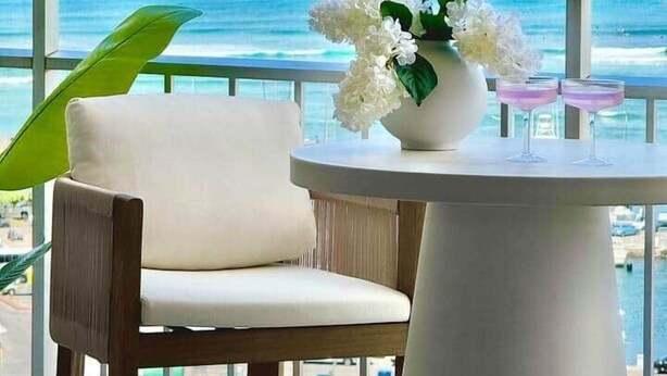 Een balkon met een witte stoel, een ronde tafel en een vaas witte bloemen.