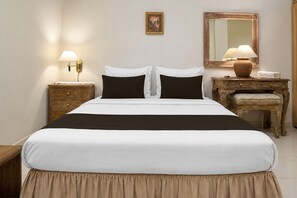 Una cama bien hecha con una colcha blanco y negro, dos mesitas de noche con lámparas, una mesita auxiliar de madera, un espejo y una pequeña mesa con un florero.