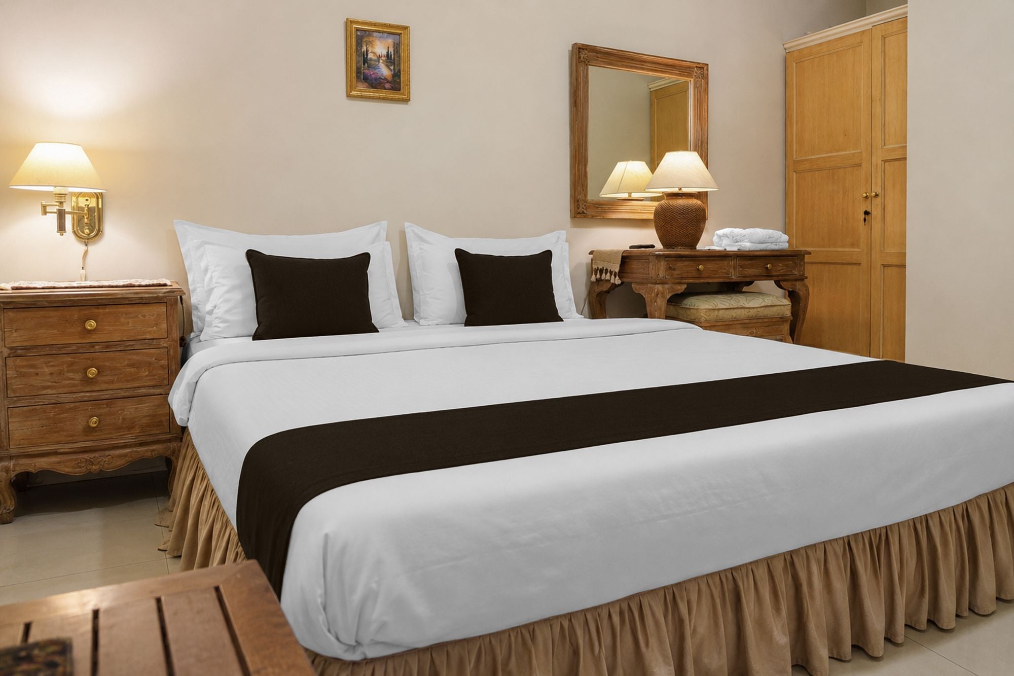 Uma cama bem arrumada com edredom branco e castanho, uma mesinha de cabeceira de madeira com abajur, um aparador de madeira com espelho e um guarda-roupa.