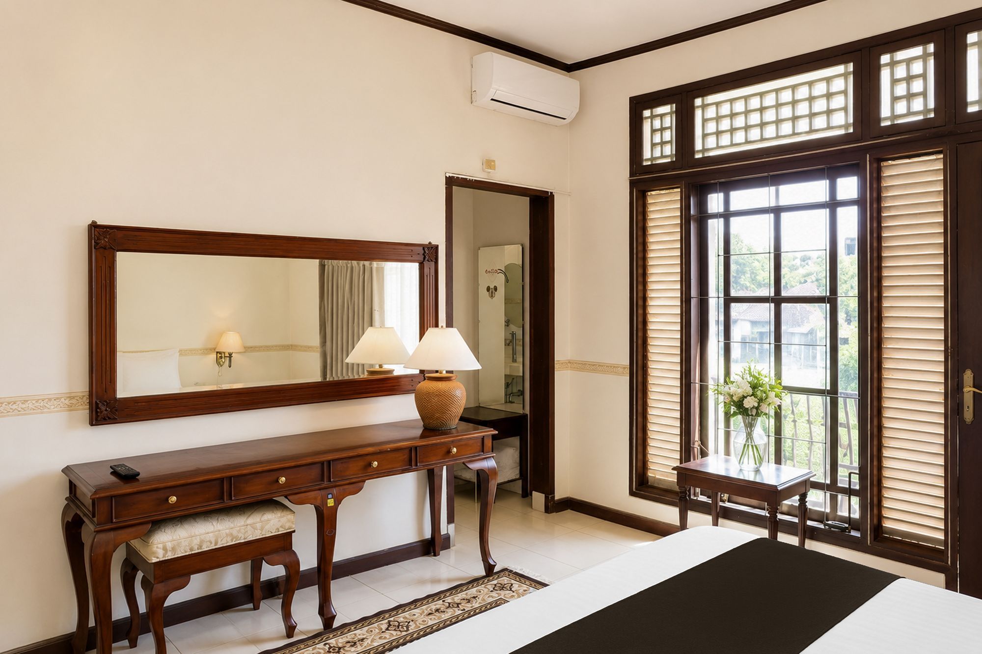 Une chambre d’hôtel avec un grand lit, une console en bois surmontée de lampes, un miroir et une fenêtre avec des stores.