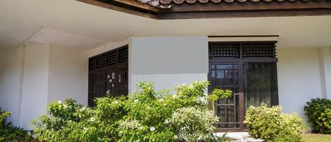 A building with a tiled roof, a balcony, and a large window. There are potted plants and a small garden area.