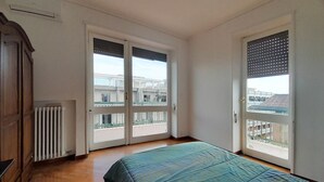 Een slaapkamer met een bed, twee deuren, een radiator en uitzicht op gebouwen.