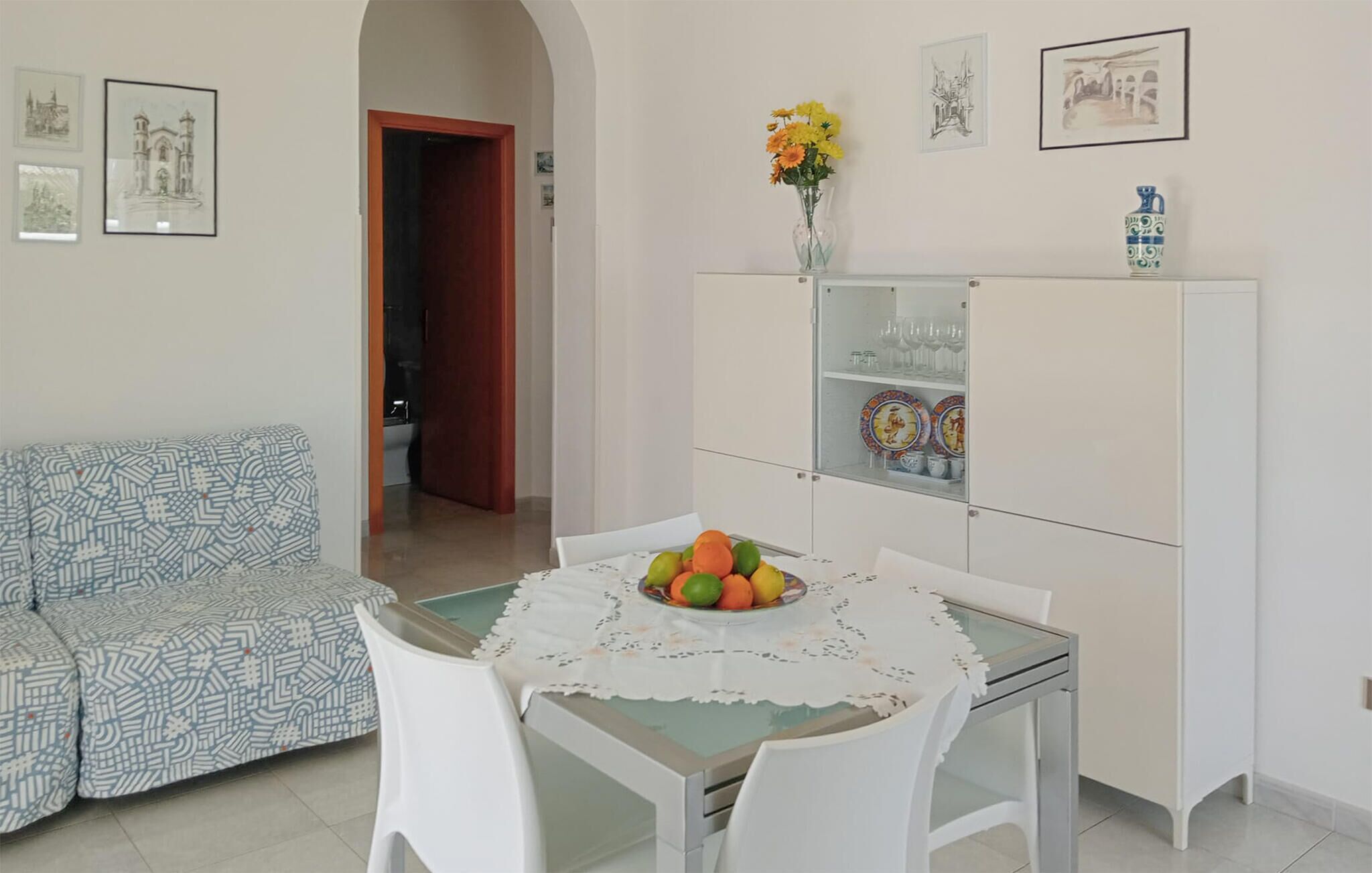 Comedor con una mesa lista para dos personas, un armario blanco con puertas de cristal y un sofá estampado.