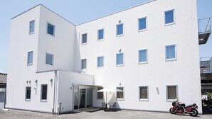 Un edificio blanco de varios pisos con fachada plana, varias ventanas y una puerta de cristal de entrada, y una motocicleta estacionada afuera.