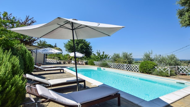 Une piscine entourée de verdure, avec des chaises longues et des parasols, sous un ciel dégagé.