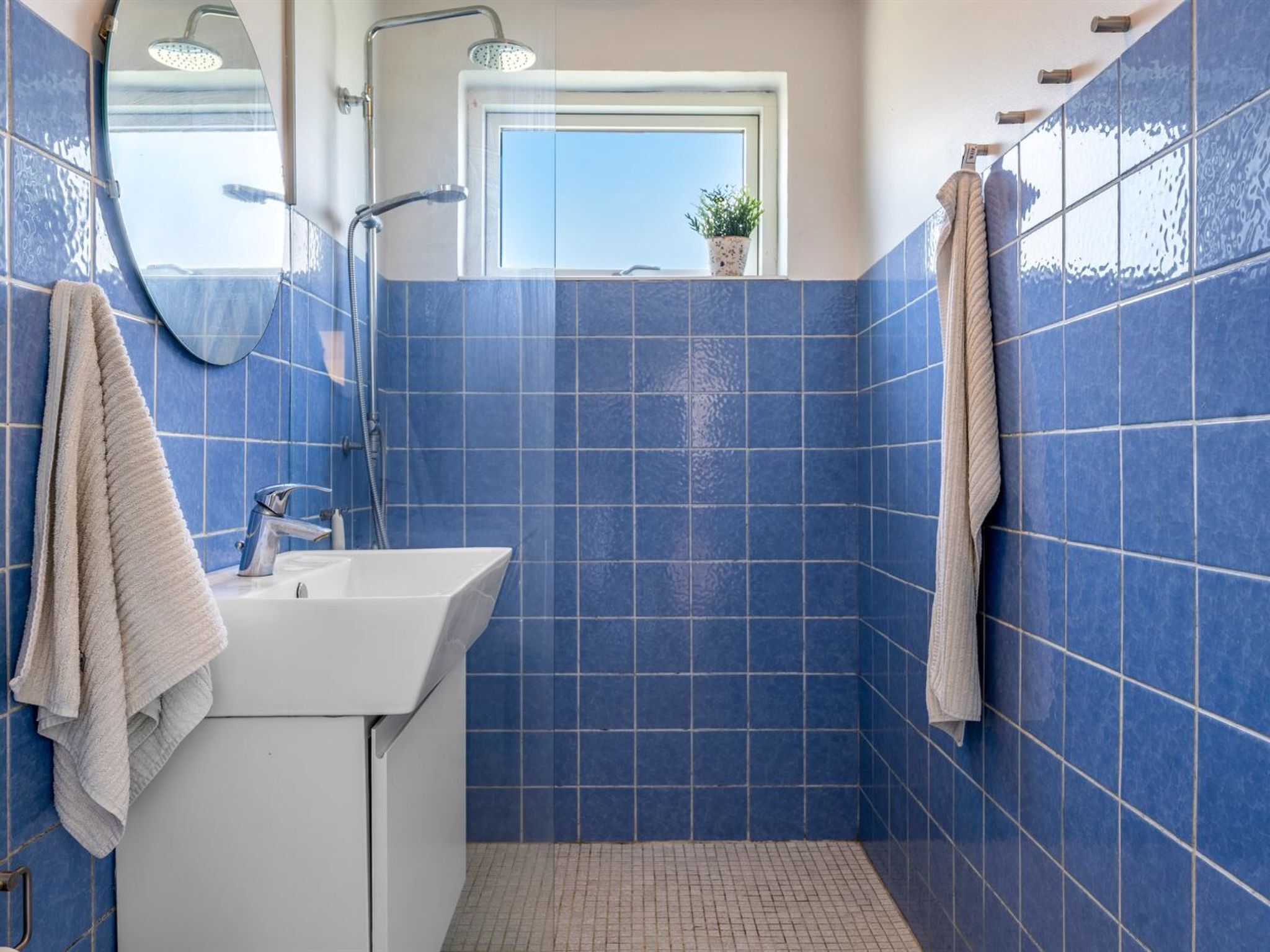 A bathroom with blue tiles, a white sink, a mirror, a shower, and a window with a plant.