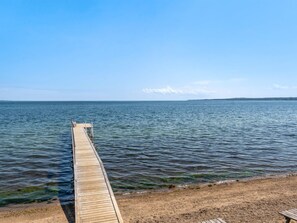 Eine hölzerne Pier, die sich in ruhiges Wasser erstreckt, ein Sandstrand und eine entfernte Küstenlinie.