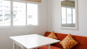 A room with a white table, an orange sofa with patterned cushions, and two windows with blinds. - Suk41 (Bangkok)