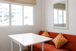 Una habitación con una mesa blanca, un sofá naranja con cojines estampados y dos ventanas con persianas.