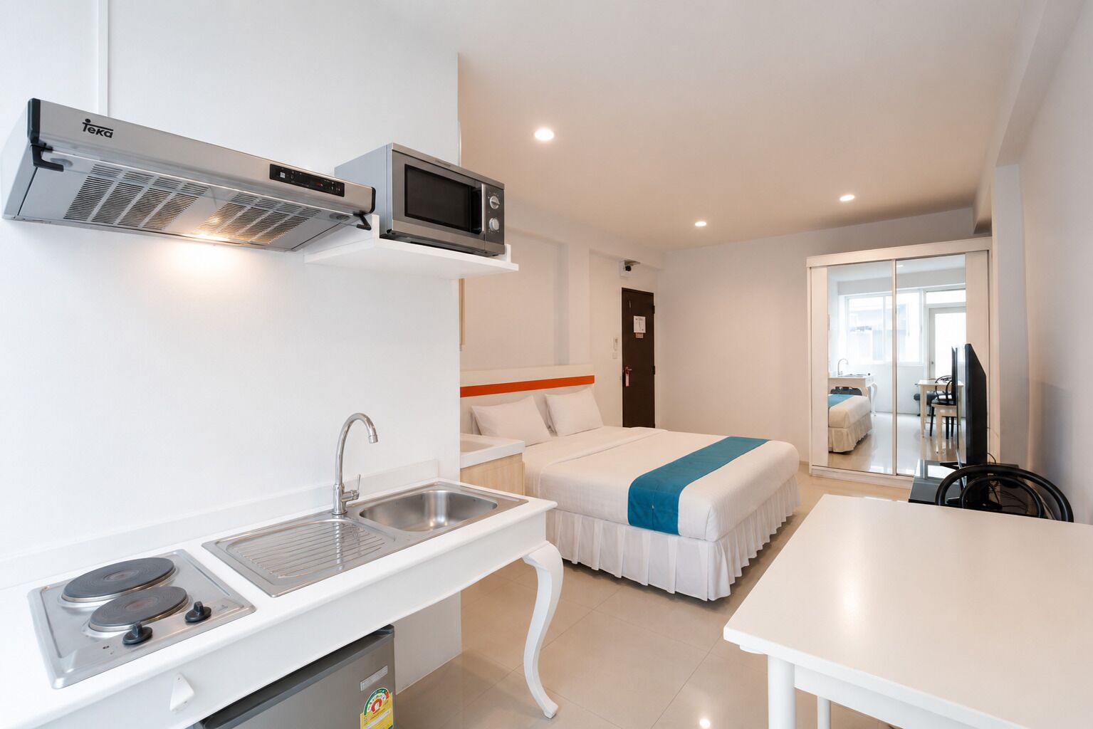 A compact hotel room with a kitchenette, a bed with a blue accent, a TV, and a bathroom.