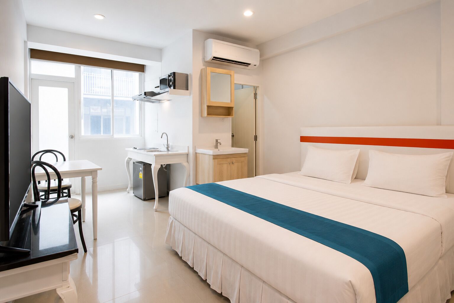 A hotel room with a bed, a desk, a chair, a TV, a sink, and an air conditioning unit.