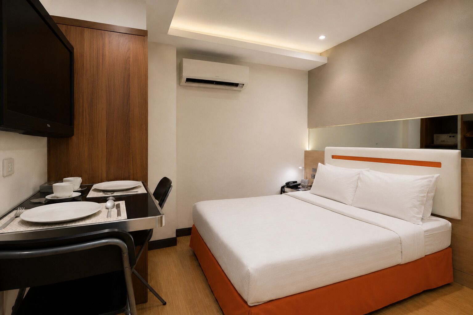 A hotel room with a bed, a desk with a chair, a flat-screen TV, and an air conditioning unit.
