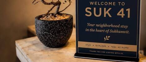 Señal de bienvenida del Sukumvit 41 Boutique Hotel, con un bonsái y una maceta texturizada.