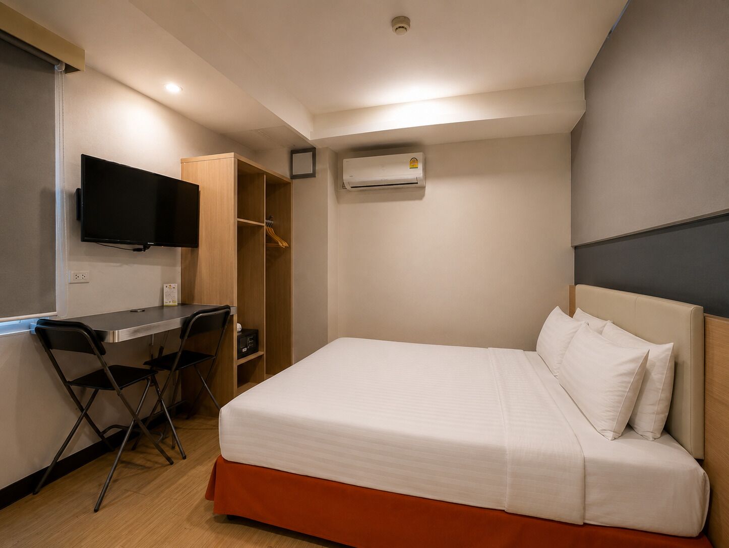 A hotel room with a bed, a desk, a chair, a TV, and an air conditioning unit.