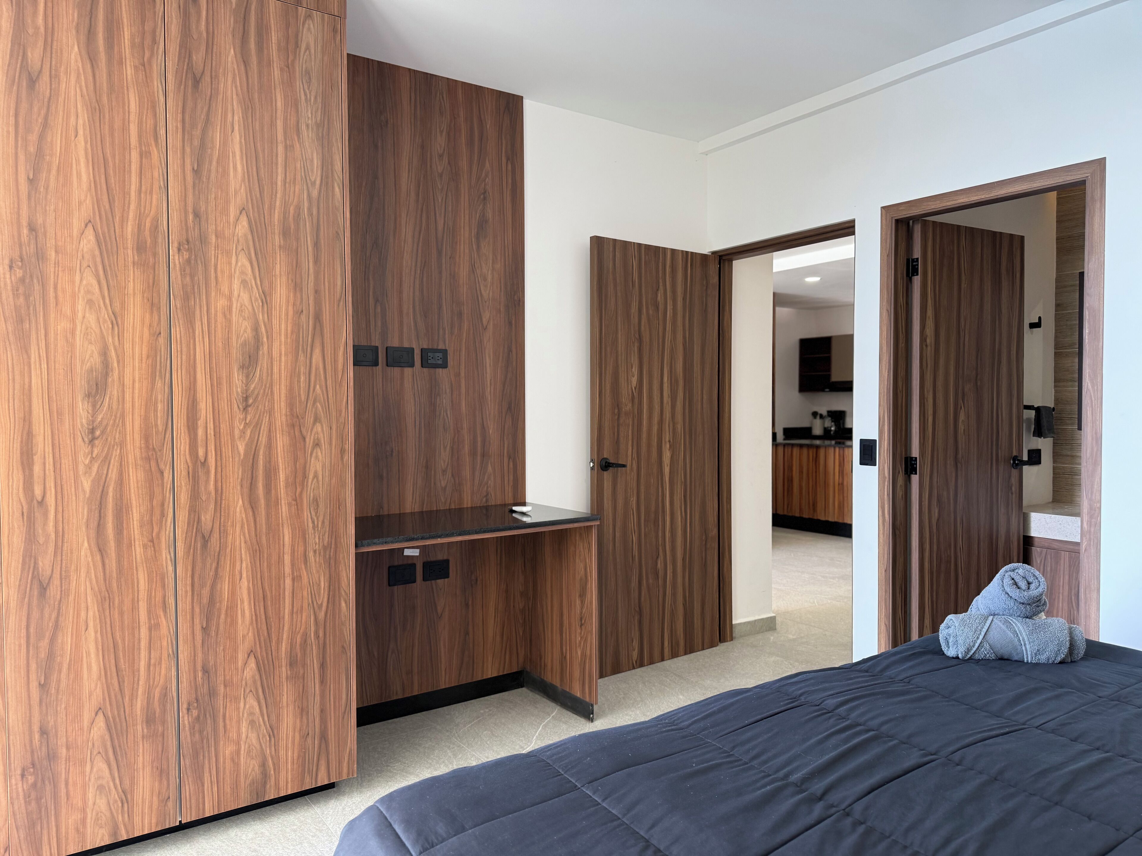 A bedroom with a bed, wooden wardrobe, and a view into the kitchen.