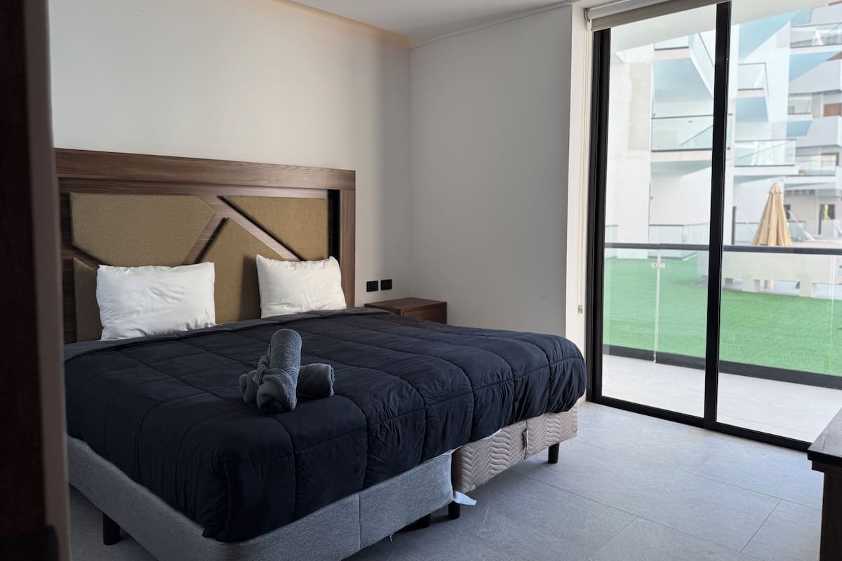 Habitación de hotel moderna con una cama grande, un cabecero de madera, una mesita de noche y vistas a un edificio y un espacio verde a través de un ventanal amplio.