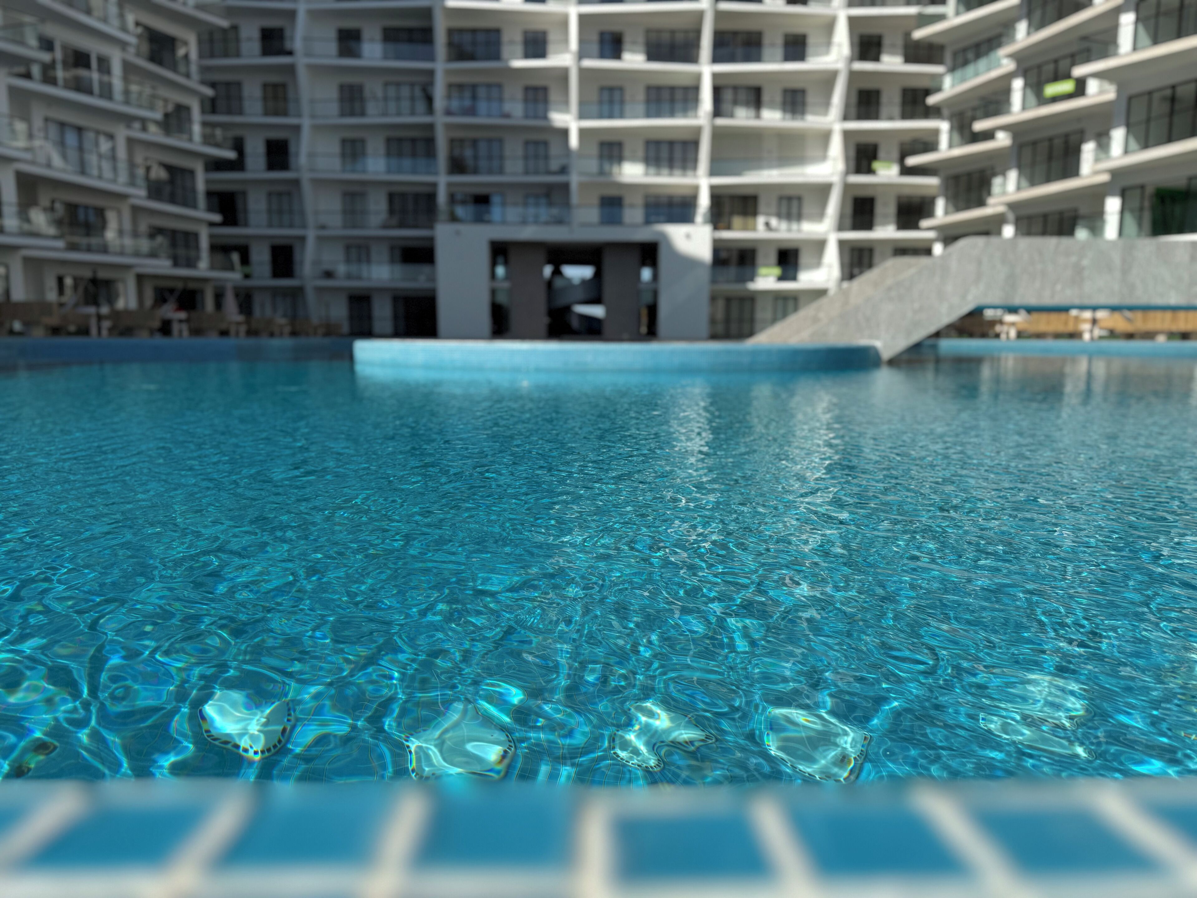 En swimmingpool med klart vand, omgivet af en moderne bygning med flere etager og altaner.