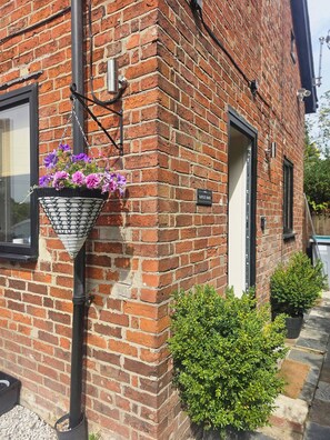 A brick building with a hanging flower basket, a door, and a small bush. - A Romantic Retreat in a Stunning Setting (Newark)
