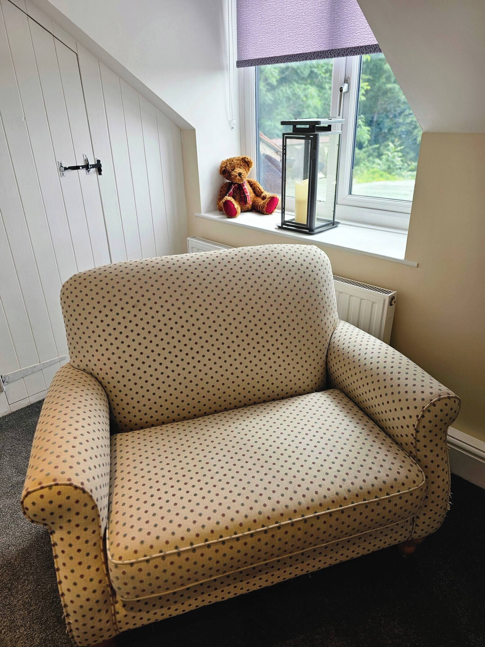 A beige polka-dotted sofa in a room with a window, a radiator, and a teddy bear on the windowsill.