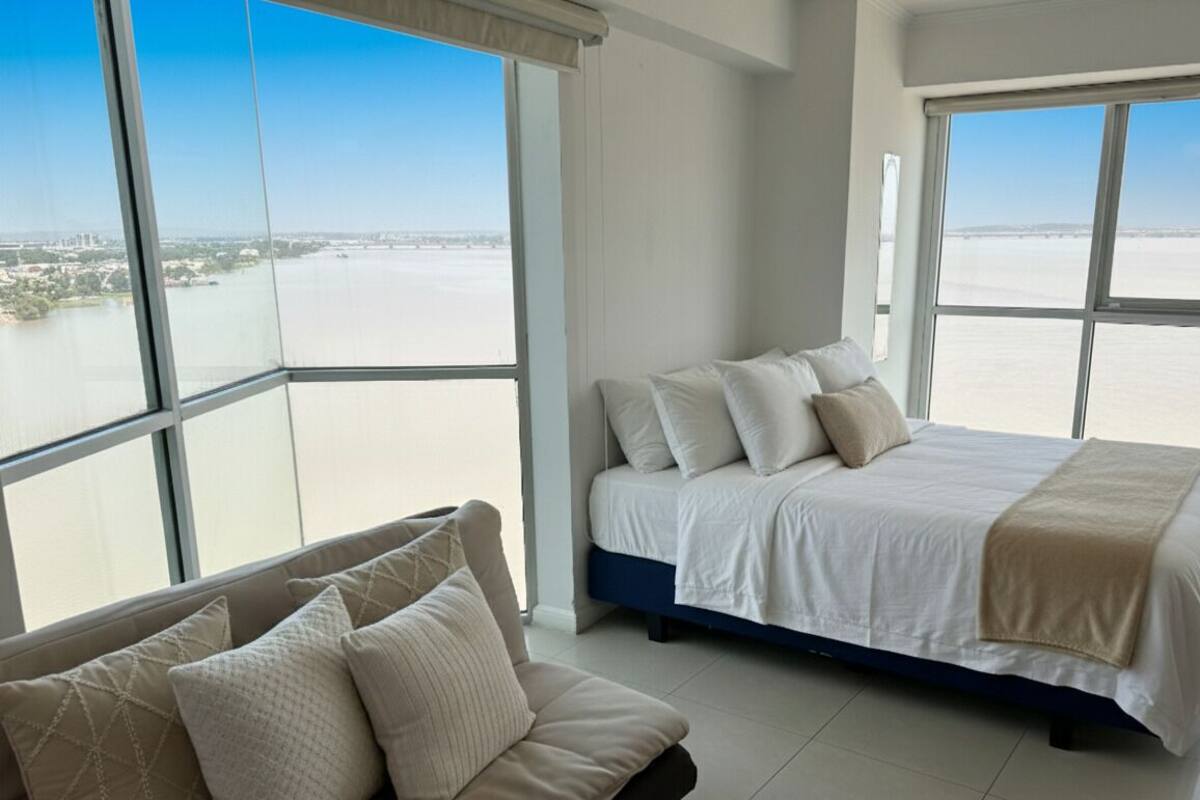Een slaapkamer met een groot bed, een slaapbank en uitzicht op een wateroppervlak door grote ramen.