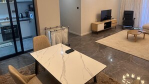 Una sala de estar moderna con una mesa de mármol, un sofá, un televisor y una cocina visible a través de un ventanal.