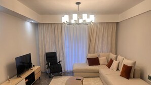 Una sala de estar moderna con un televisor de pantalla plana, una silla de oficina negra, un sofá beige con cojines marrones, una pequeña mesa de centro de madera y una araña.