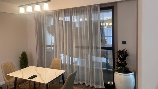 Una habitación moderna con una mesa de comedor, sillas, una planta grande en maceta y vistas a través de cortinas traslúcidas.