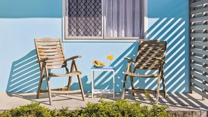 Two wooden chairs with a table between them, a window with a mesh screen and curtains, and a blue wall with a white lattice structure. - Little Banksia  - Pet-Friendly in the Heart of Yamba - Holiday Yamba (Yamba)