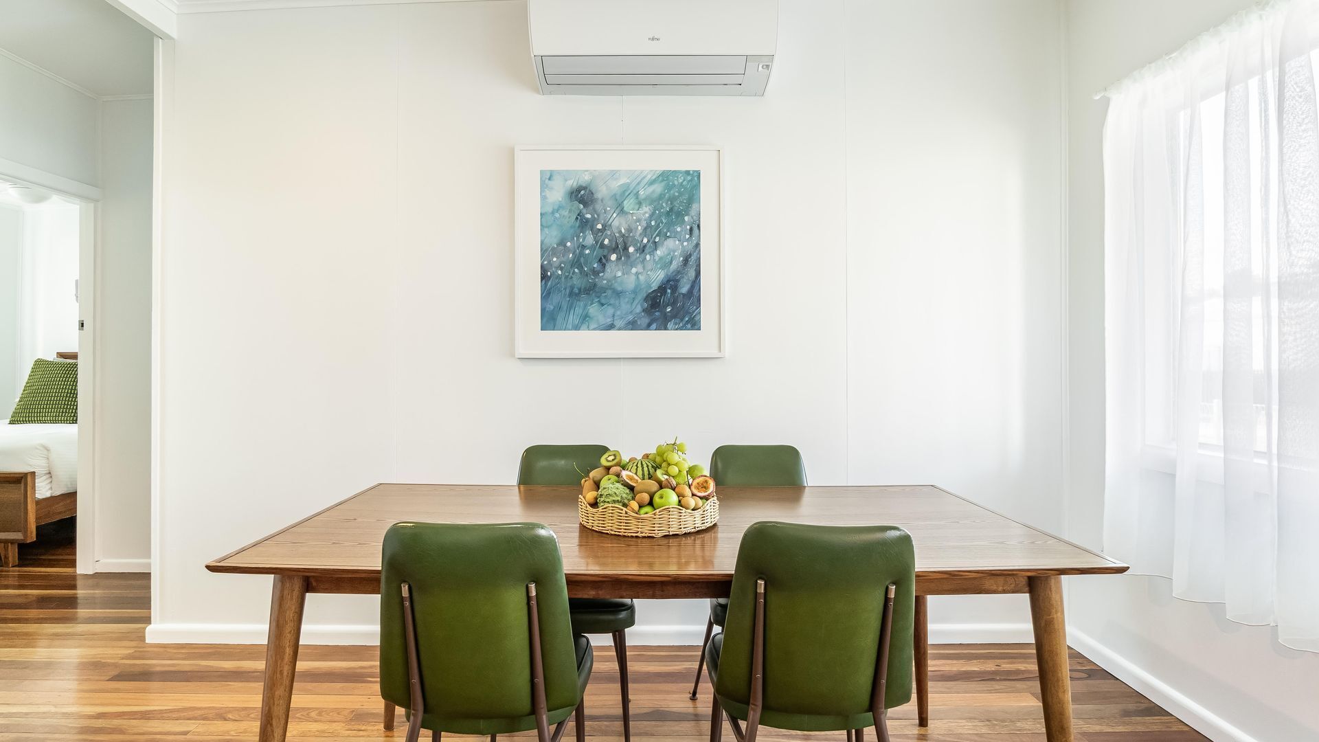 A dining area with a wooden table, four green chairs, a fruit basket, and a blue abstract painting on the wall.