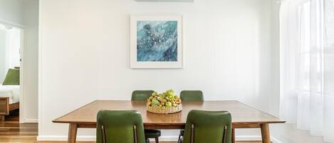 A dining area with a wooden table, four green chairs, a fruit basket, and a blue abstract painting on the wall.