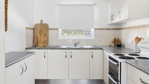 A modern kitchen with white cabinets, a wooden cutting board, a loaf of bread, a window with a decorative grid, and a stove. - Little Banksia  - Pet-Friendly in the Heart of Yamba - Holiday Yamba (Yamba)